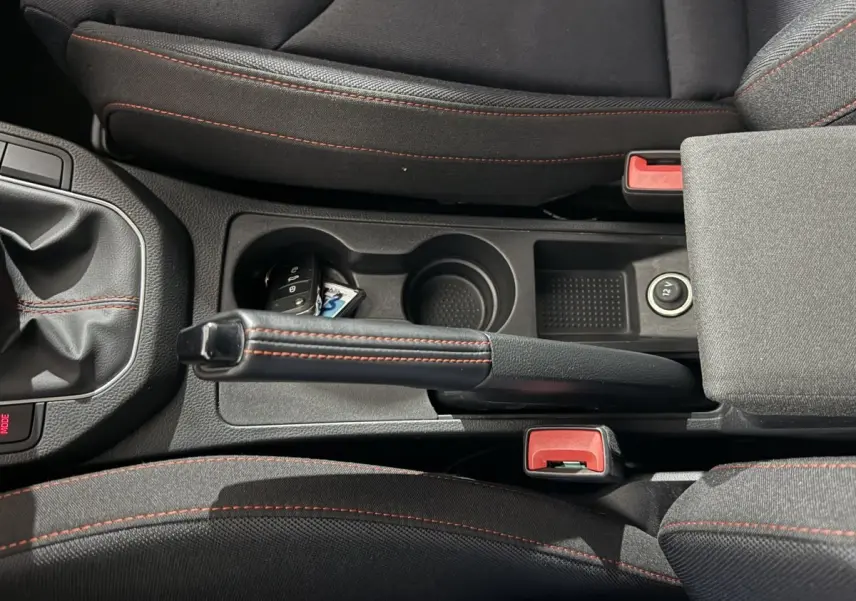 Vue plongeante sur la console centrale noire avec frein à main cuir et coutures rouges, clé et prise 12V visibles.