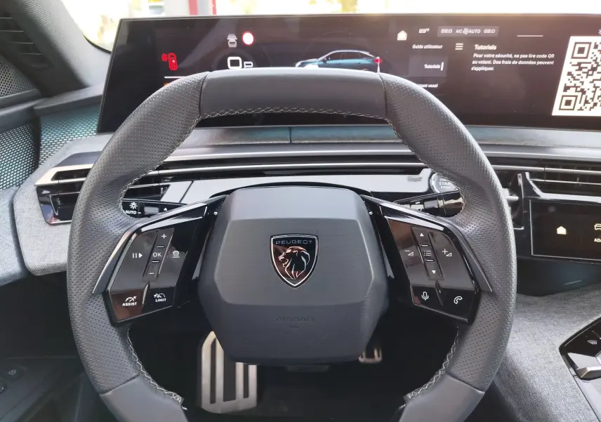 Vue rapprochée du volant multifonction noir avec logo Peugeot, tableau de bord numérique et commandes tactiles visibles.