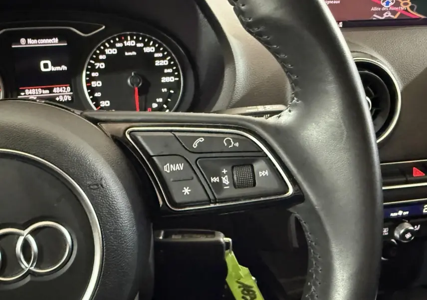 Vue rapprochée du volant cuir noir d’Audi A3 2018 avec commandes audio et tableau de bord digital.