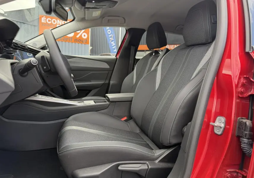 Intérieur côté conducteur d’une Peugeot 308 SW rouge, sièges noirs avec surpiqûres bleues et accoudoir central visible.