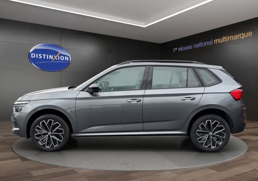 Vue latérale droite d'un Skoda Kamiq gris foncé avec jantes bi-ton et barres de toit noires en showroom.