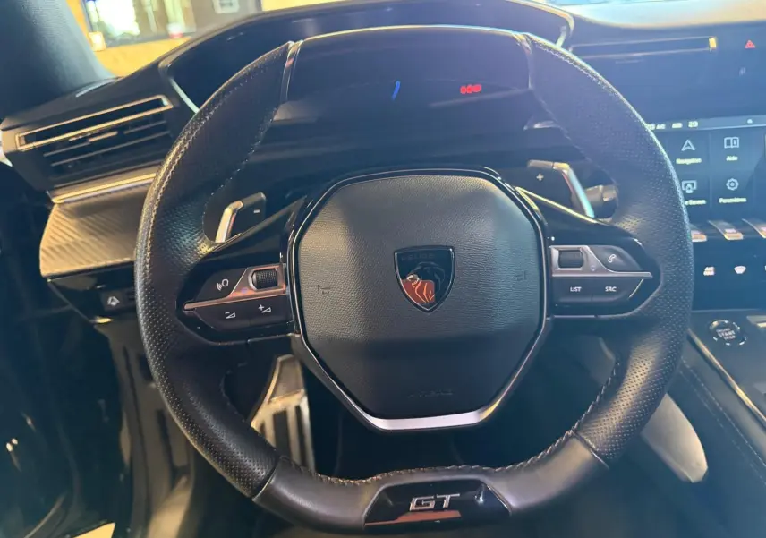 Volant cuir perforé noir avec badge Peugeot et sigle GT, tableau de bord et commandes tactiles en arrière-plan.