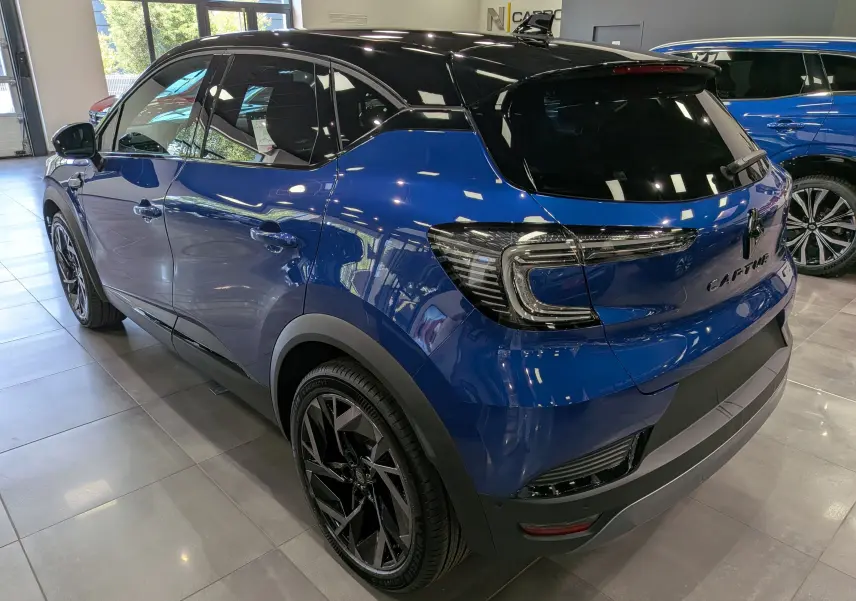 Renault Captur E-Tech hybride 2026 bleu Iron avec toit noir, vue 3/4 arrière droit en intérieur showroom.
