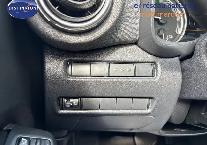 Gros plan sur le commodo d’éclairage à gauche du volant du Nissan Juke Hybrid gris, avec tableau de bord numérique en arrière-plan.