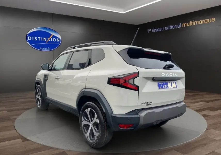 Vue 3/4 arrière droite du Dacia Duster beige 2025 avec barres de toit noires et jantes alliage visibles.