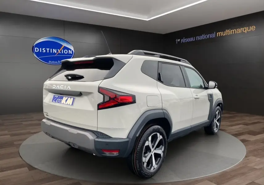 Vue 3/4 arrière droite du Dacia Duster beige 2025 avec jantes alliage et barres de toit noires.