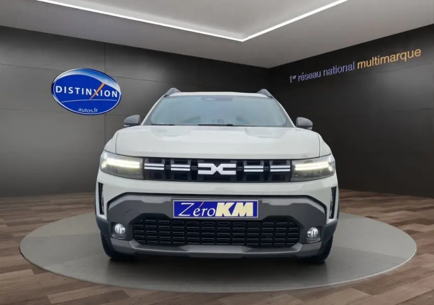 Vue avant d'un Dacia Duster beige 2025 avec calandre noire et phares LED allumés en showroom.