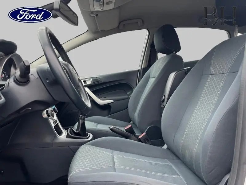Vue intérieure côté conducteur de la Ford Fiesta 2012, sellerie tissu gris avec volant cuir et levier de vitesses manuel.