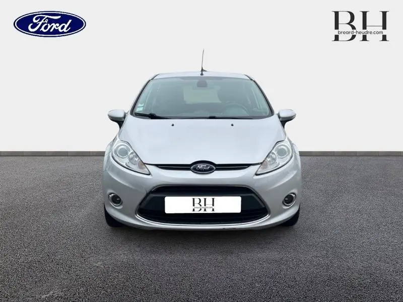 Vue avant d'une Ford Fiesta gris lunaire 2012 avec calandre chromée et phares halogènes allumés.