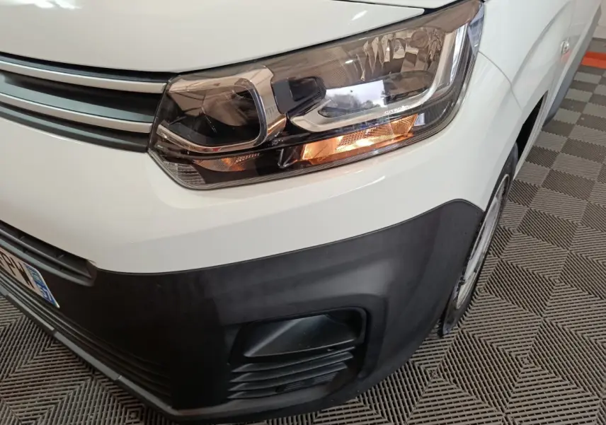 Gros plan sur le phare avant droit et la calandre noire d’un Citroën Berlingo blanc, vue en légère plongée.