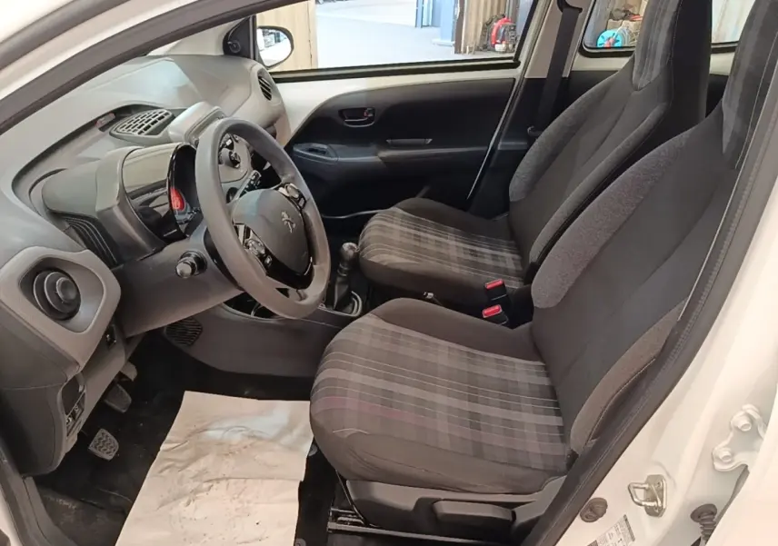 Vue intérieure côté conducteur de la Peugeot 108 blanche, montrant les sièges avant à motifs carreaux et le volant multifonctions.