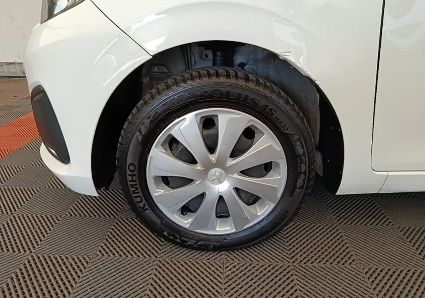 Gros plan sur la roue avant gauche d'une Peugeot 108 blanche, avec enjoliveur argenté et carrosserie légèrement froissée.
