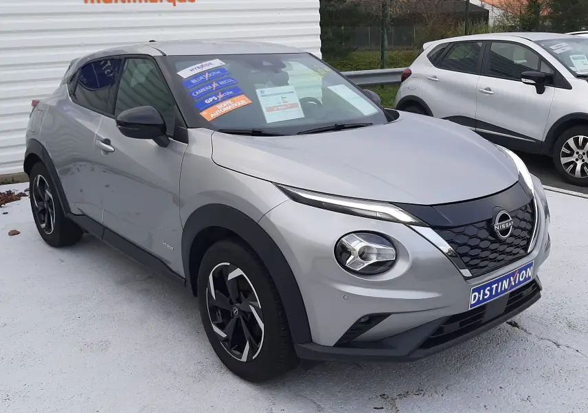 Nissan Juke 1.6 HEV gris perle métal vu en 3/4 avant droit, avec jantes noires et stickers promotionnels sur le pare-brise.