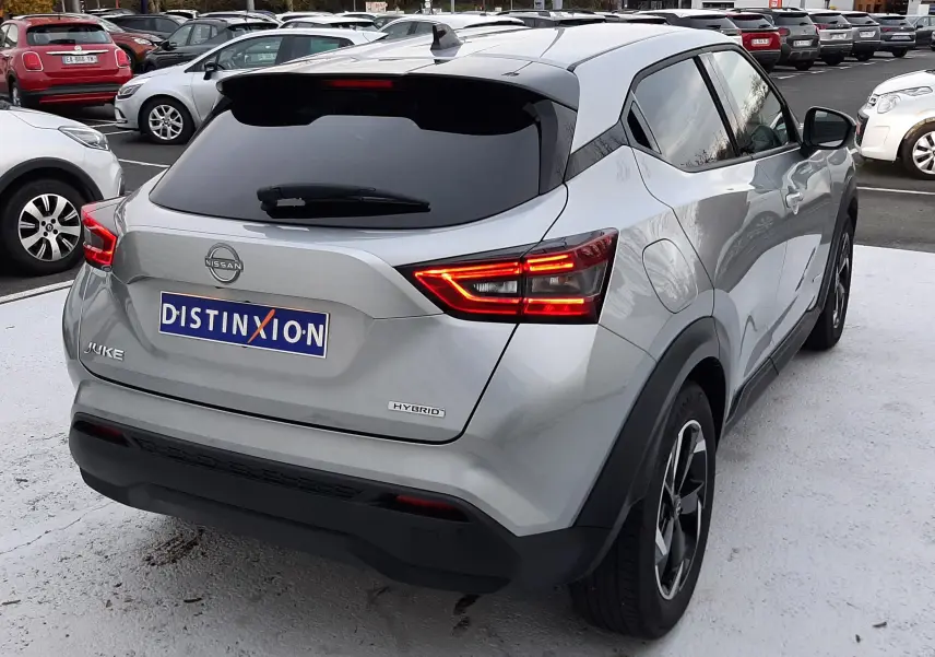 Vue 3/4 arrière droite d'un Nissan Juke gris perle métal hybride 2024 avec feux arrière allumés sur parking.