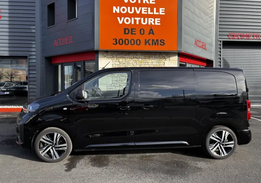 Profil côté gauche d’un FIAT Scudo Fourgon noir 2025 garé devant un bâtiment avec enseigne orange.