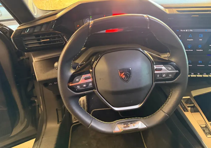 Vue intérieure centrée sur le volant noir perforé avec logo Peugeot et badge GT, planche de bord et écran tactile visibles.
