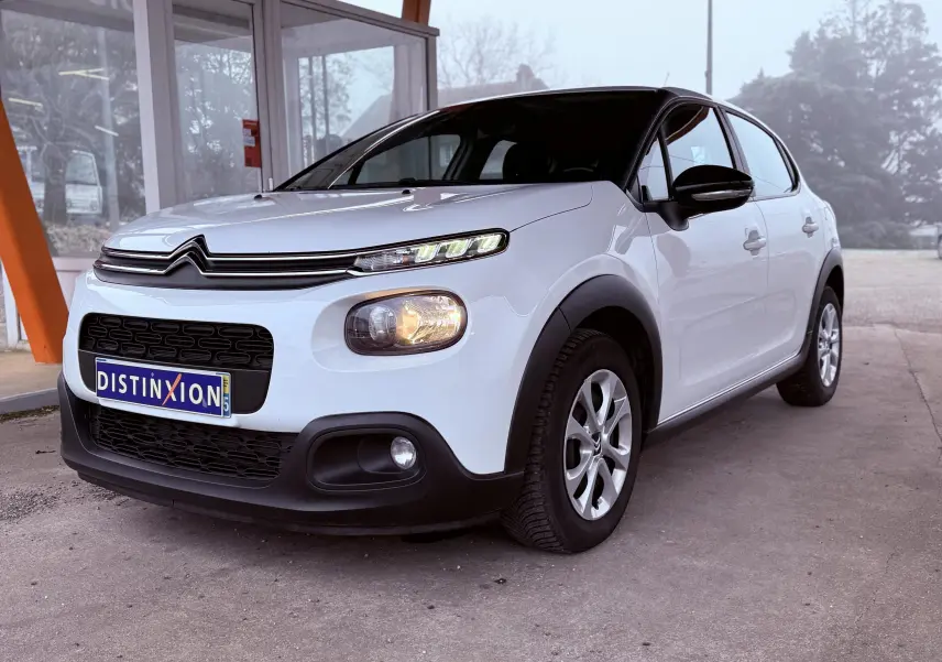 Citroën C3 blanc vue 3/4 avant droit, avec calandre noire et phares allumés sous abri.