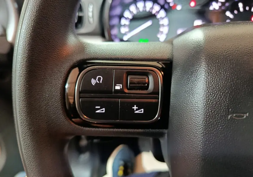 Gros plan sur les commandes gauche du volant multifonctions noir du Citroën C3 2024, avec tableau de bord flou en arrière-plan.