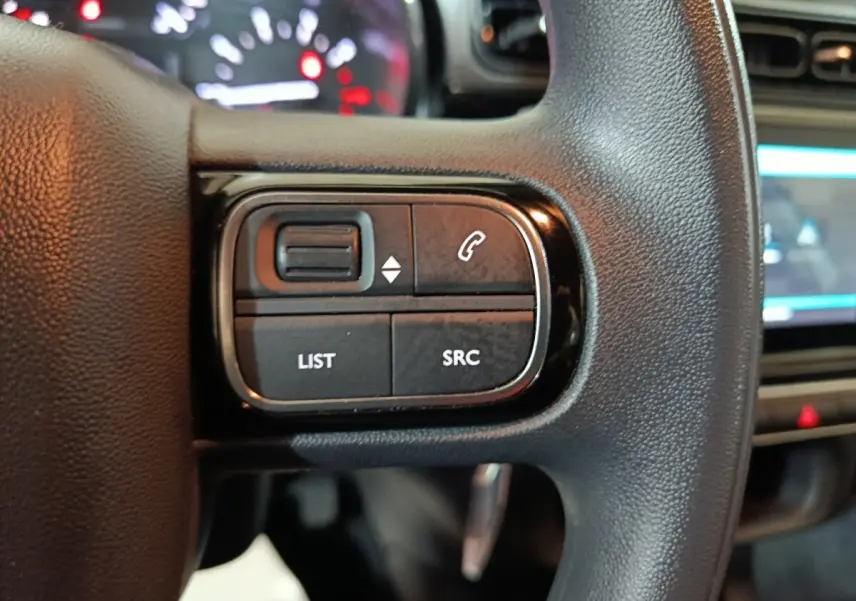 Gros plan sur les commandes gauche du volant multifonctions noir de la Citroën C3 2024, avec tableau de bord flou en arrière-plan.