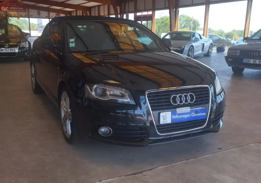 Audi A3 Cabriolet noir vue 3/4 avant droit dans un showroom avec plusieurs autres véhicules exposés.