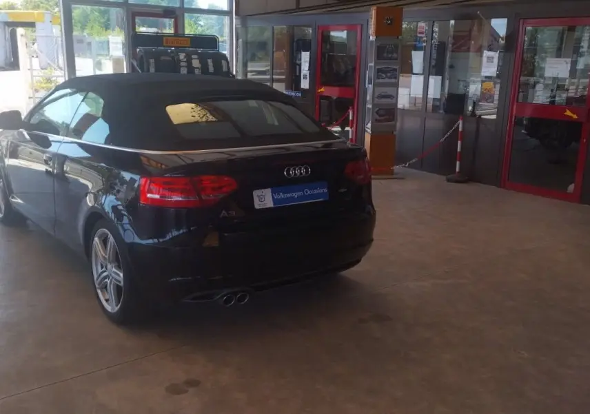 Audi A3 Cabriolet noir vue 3/4 arrière droit, capote fermée, garée dans un garage lumineux avec portes vitrées rouges.
