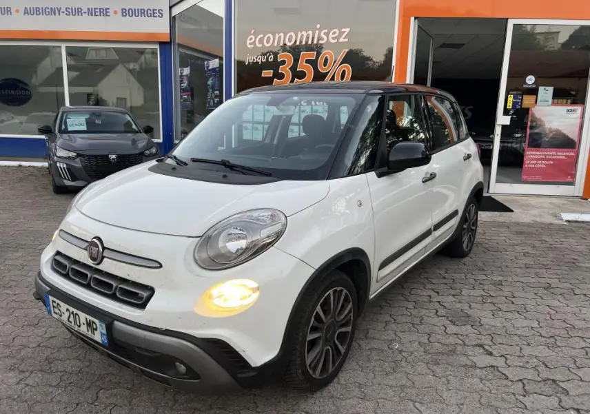 Vue 3/4 avant d'une Fiat 500L blanche avec toit noir et feux diurnes allumés devant un showroom.