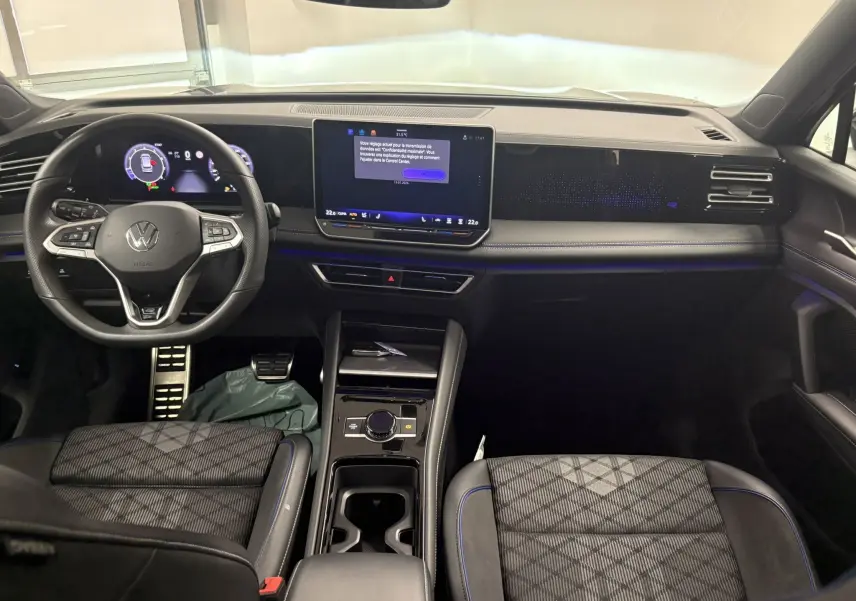Vue intérieure du tableau de bord et volant du Volkswagen Tiguan 2025, finition R-Line avec écran tactile et sièges tissu noir à surpiqûres bleues.