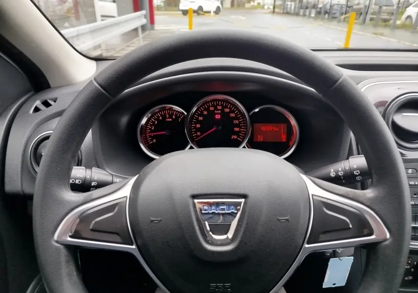 Vue rapprochée du volant et du tableau de bord d'une Dacia Sandero blanche, avec compteurs et indicateurs rouges visibles.