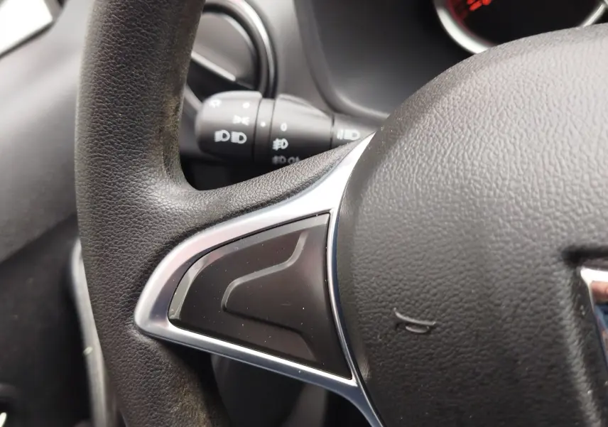 Gros plan sur la partie gauche du volant noir texturé d'une Dacia Sandero blanc glacier, avec commandes intégrées visibles.