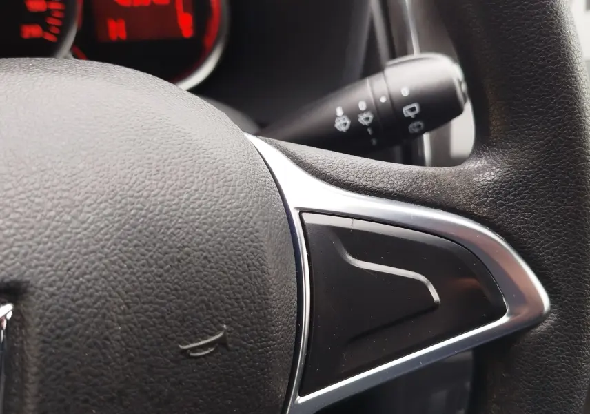 Gros plan sur le volant en cuir noir de la Dacia Sandero blanc glacier, avec commandes intégrées et tableau de bord rouge en arrière-plan.