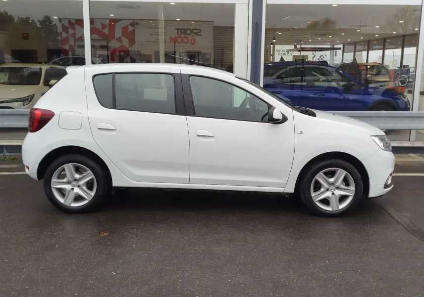 Profil latéral droit d'une Dacia Sandero blanc glacier de 2019, compacte et cinq portes, stationnée devant une vitrine.