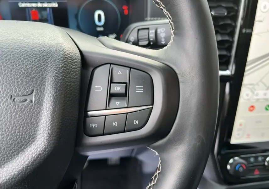Gros plan sur les commandes au volant cuir noir du Ford Ranger 2026, avec écran tactile et tableau de bord numérique en arrière-plan.