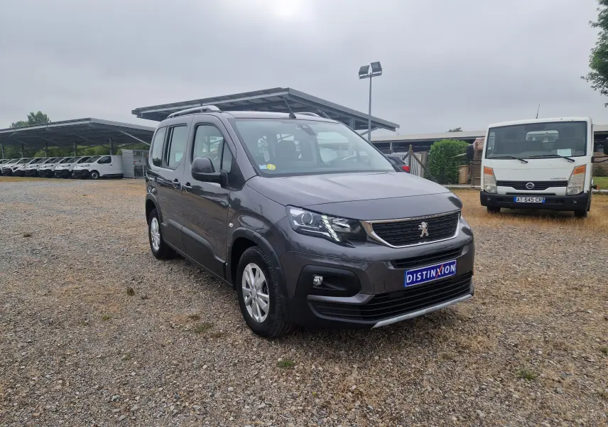 Peugeot Rifter gris platinium en 3/4 avant droit sur un sol gravillonné avec plusieurs véhicules en arrière-plan.