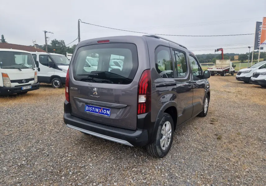 Vue 3/4 arrière droite d'un Peugeot Rifter gris platinium avec hayon et feux arrière verticaux, stationné sur un sol gravillonné.