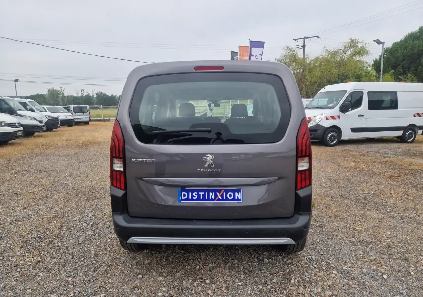 Vue arrière du Peugeot Rifter gris platinium 2019, avec feux arrière verticaux et hayon vitré sur un terrain gravillonné.