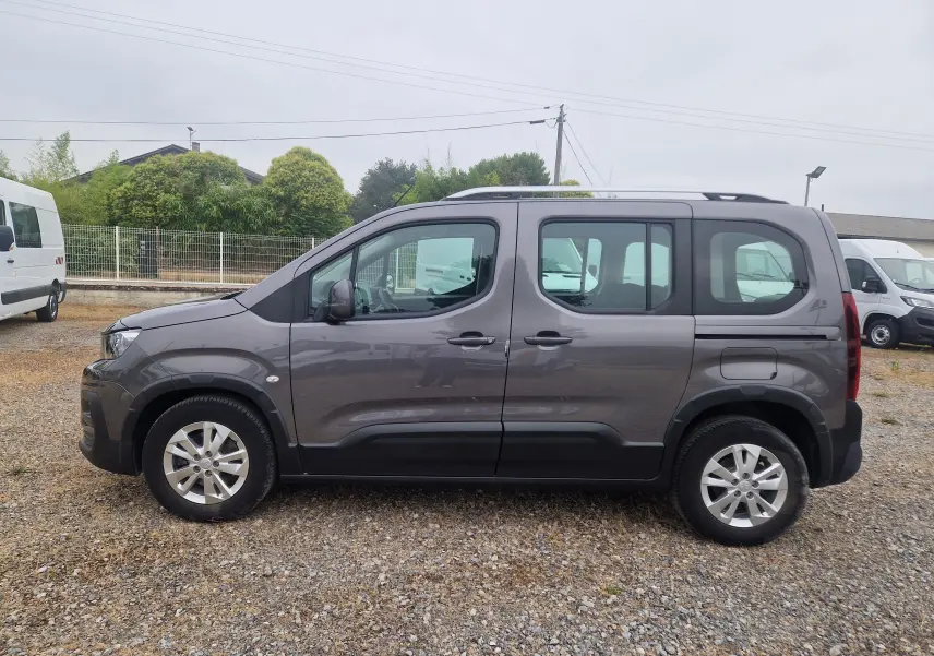 Vue 3/4 arrière droite d'un Peugeot Rifter gris platinium garé sur un sol gravillonné avec plusieurs véhicules autour.