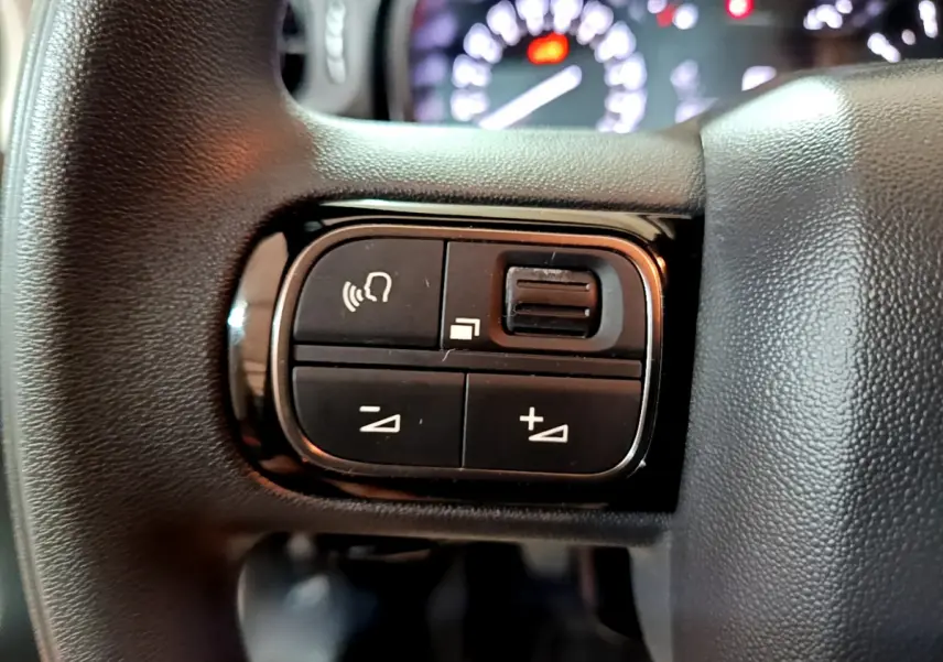 Gros plan sur les commandes multifonctions noires à gauche du volant du Citroën C3 2022 beige, avec tableau de bord flou en arrière-plan.