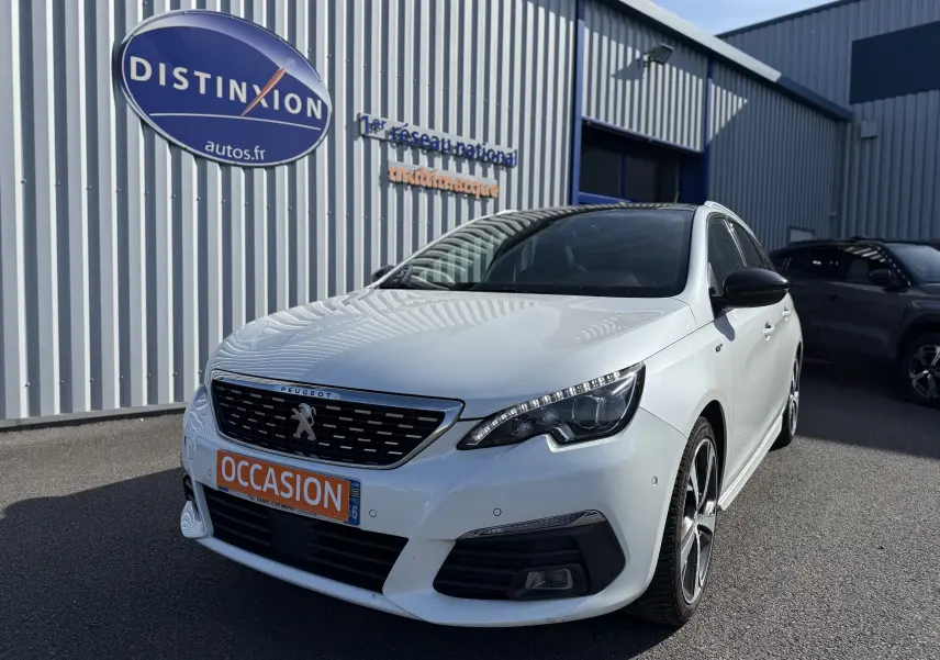 Peugeot 308 SW blanc nacré vue 3/4 avant droit, avec calandre noire et plaque orange "OCCASION" devant un bâtiment industriel.