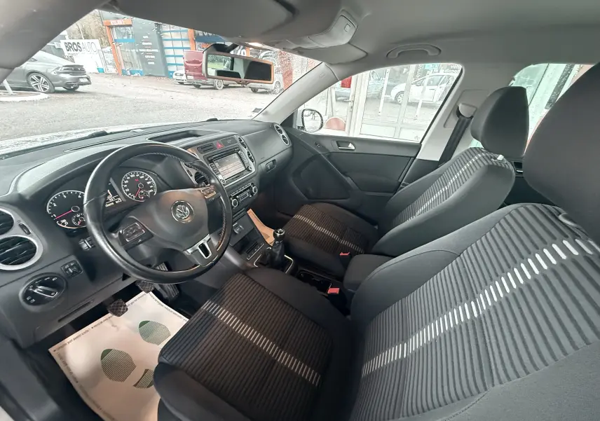 Vue intérieure côté conducteur du Volkswagen Tiguan blanc de 2010, montrant le volant cuir, tableau de bord et sièges tissu noir rayé.
