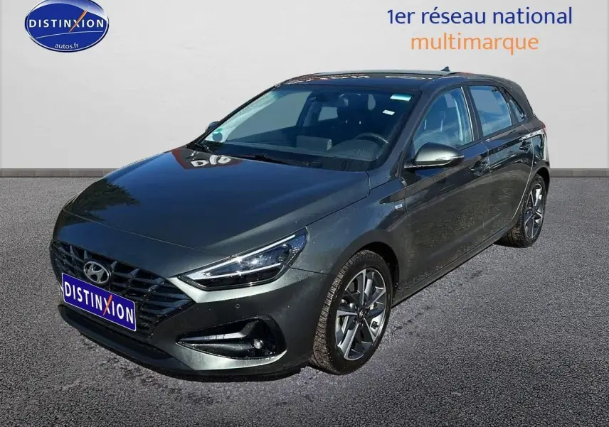Hyundai i30 gris argent vue 3/4 avant droit avec jantes alu et phares LED allumés sur parking.