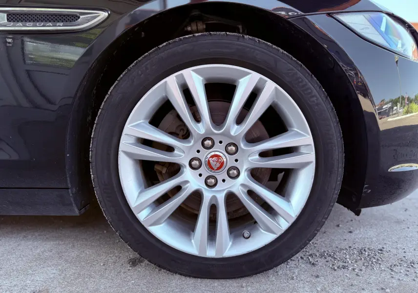 Gros plan sur la roue avant gauche d'une Jaguar XE noire, mettant en valeur la jante argentée et l'emblème Jaguar rouge.