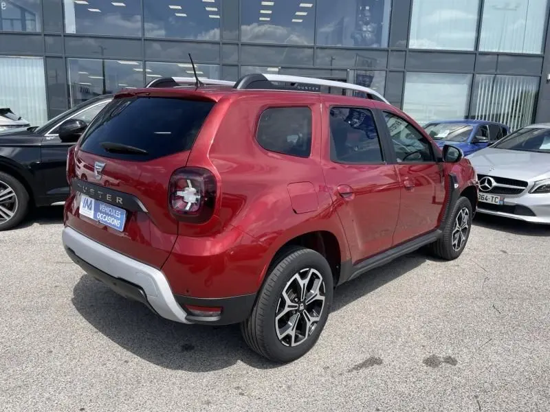 Vue 3/4 arrière droite d'un Dacia Duster TCE 150 Prestige 4x2 rouge métal avec jantes alliage et barres de toit.