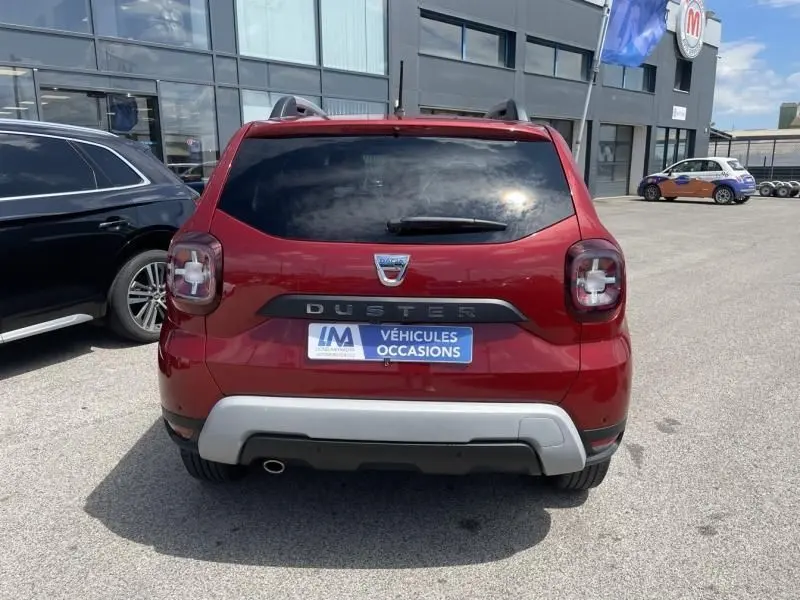 Vue arrière d'un Dacia Duster TCE 150 Prestige 4x2 rouge métallisé garé devant un bâtiment commercial.