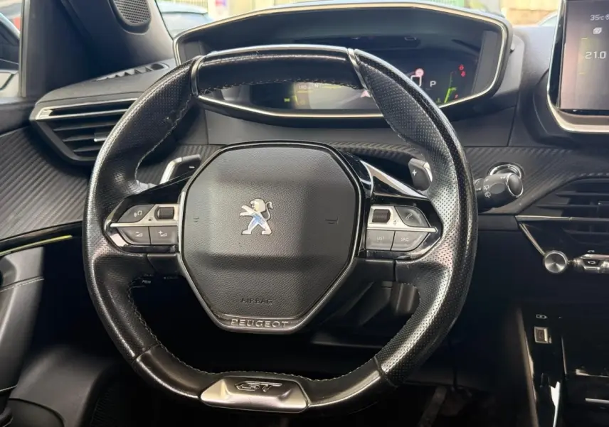 Vue rapprochée du volant noir perforé avec logo Peugeot et badge GT, tableau de bord et écran digital en arrière-plan.