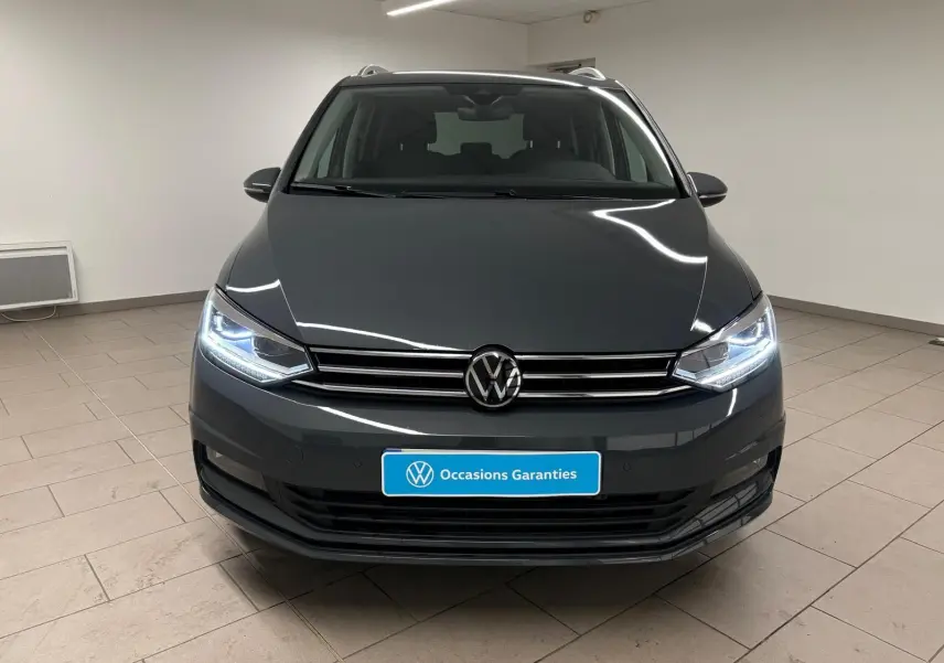 Vue frontale d'un Volkswagen Touran gris dauphin avec phares allumés dans un showroom intérieur.