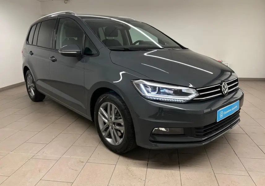 Vue 3/4 avant gauche d’un Volkswagen Touran 2025 gris dauphin avec phares allumés dans un showroom.