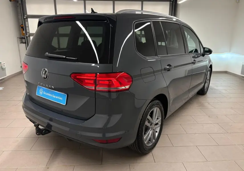Vue 3/4 arrière droite d’un Volkswagen Touran gris dauphin avec attelage escamotable dans un garage lumineux.