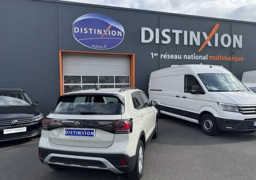 Vue arrière d'un Volkswagen T-Cross gris Ascot garé devant un bâtiment Distinxion avec un utilitaire blanc à côté.