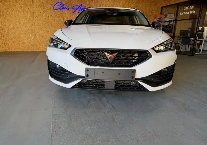 Vue frontale d'une CUPRA LEON ST blanche 2024 avec calandre noire et logo cuivre, dans un garage intérieur.