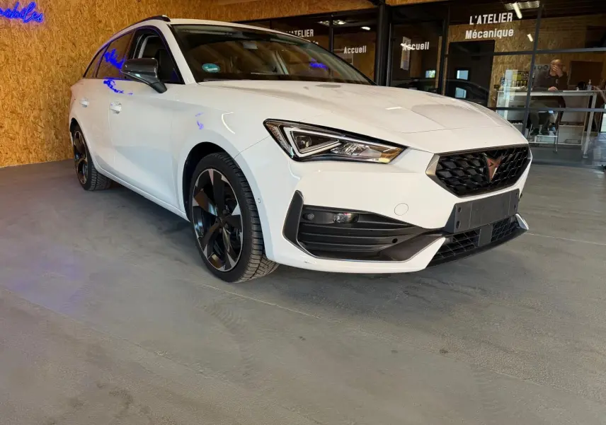 Vue 3/4 avant droit d'une CUPRA LEON ST blanche 2024 avec jantes noires et calandre noire dans un garage.